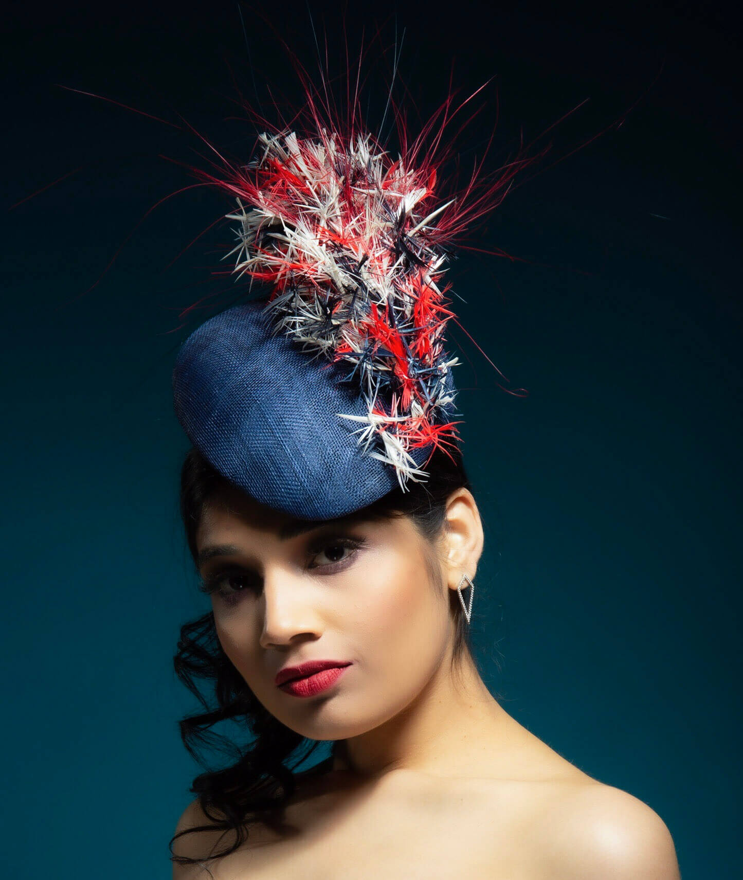 Woman wearing a unique blue fascinator adorned with red and white feathers against a dark background. | Sarah Ross Millinery