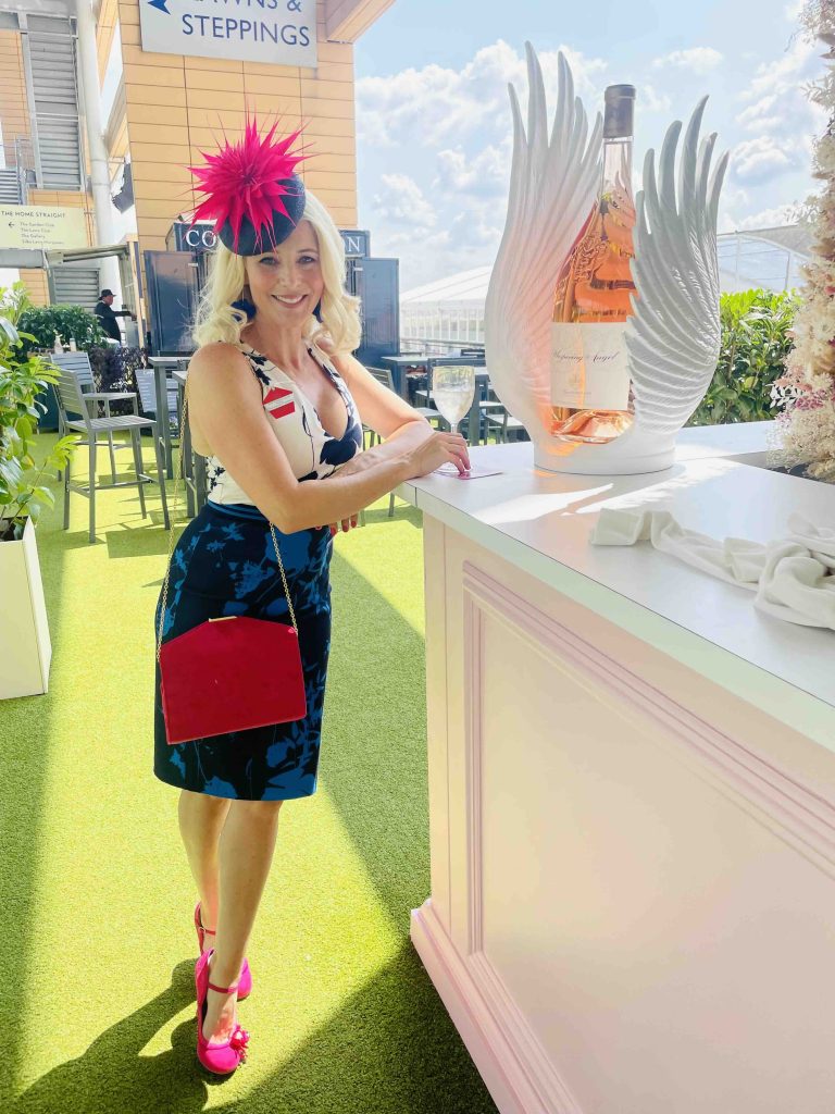 A woman in a colorful dress and pink heels stands by a bar with a large bottle of rosé wine. | Sarah Ross Millinery