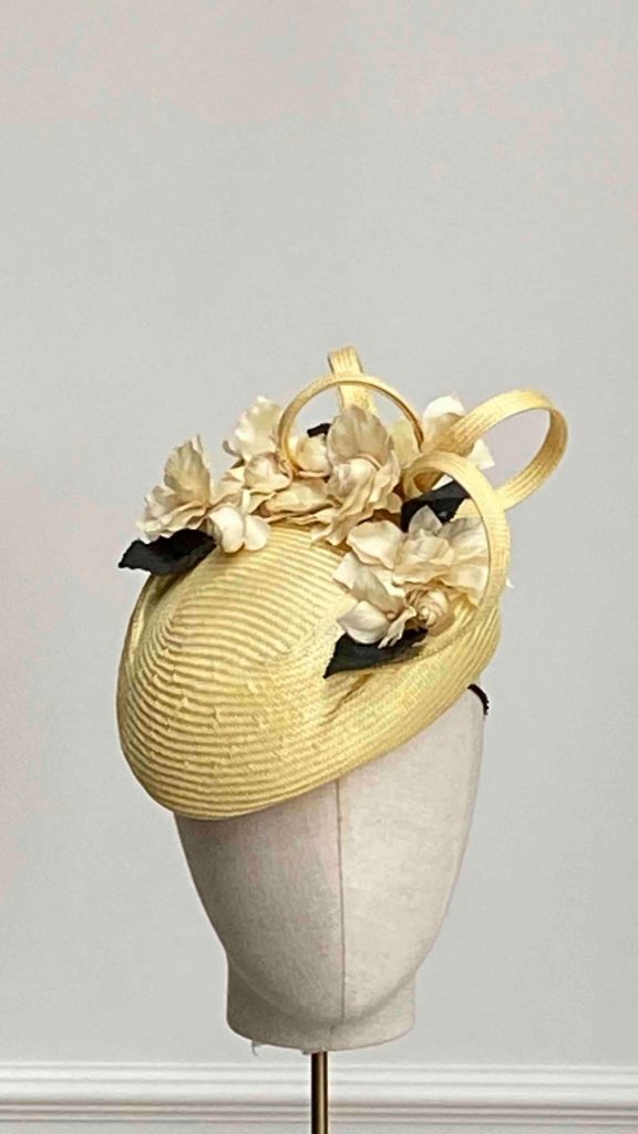 A cream-colored hat with ribbon and floral decorations displayed on a mannequin head against a plain background. | Sarah Ross Millinery