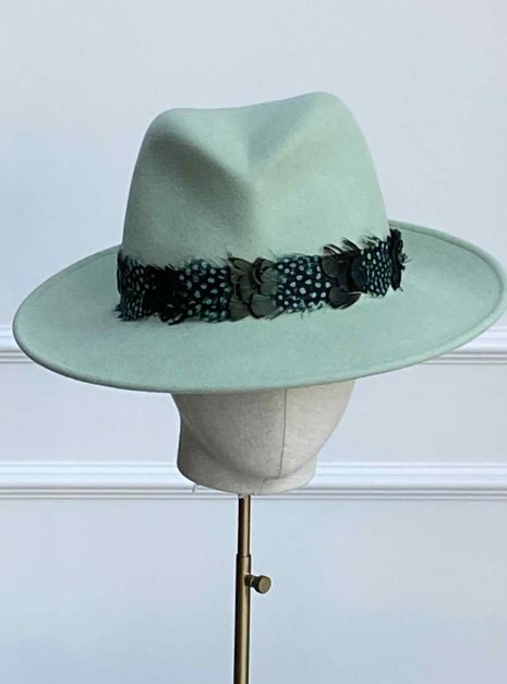 A light green fedora hat on a stand, featuring a dark patterned band with decorative elements, set against a plain background. | Sarah Ross Millinery