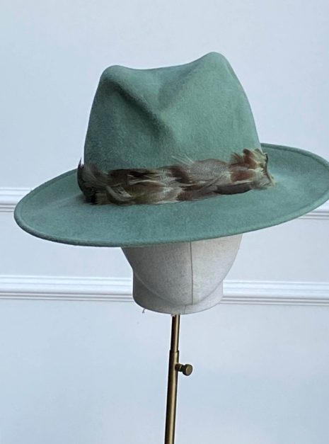 A green fedora hat with a feather band displayed on a stand against a light background. | Sarah Ross Millinery