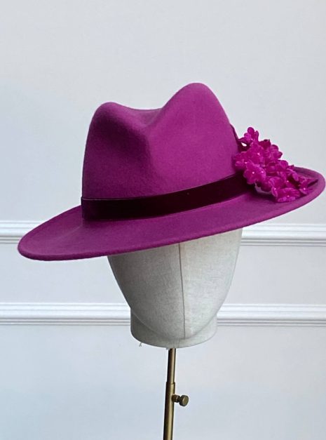 A magenta fedora hat with flower decorations on the side, displayed on a mannequin head with a stand against a plain background. | Sarah Ross Millinery