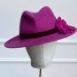 A magenta fedora hat with flower decorations on the side, displayed on a mannequin head with a stand against a plain background. | Sarah Ross Millinery