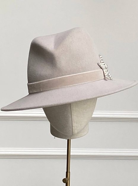 A light gray fedora hat with a wide brim and a ribbon band, adorned with a metallic feather, displayed on a mannequin head with a neutral background. | Sarah Ross Millinery