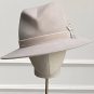 A light gray fedora hat with a wide brim and a ribbon band, adorned with a metallic feather, displayed on a mannequin head with a neutral background. | Sarah Ross Millinery