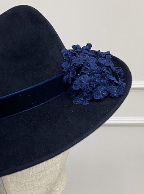 IMG_1940 A dark blue felt hat with a wide brim, featuring a navy ribbon and a cluster of blue floral appliques on one side. | Sarah Ross Millinery