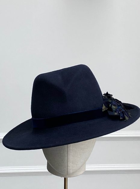A dark blue wide-brimmed hat displayed on a mannequin head, featuring a subtle floral decoration on one side. | Sarah Ross Millinery