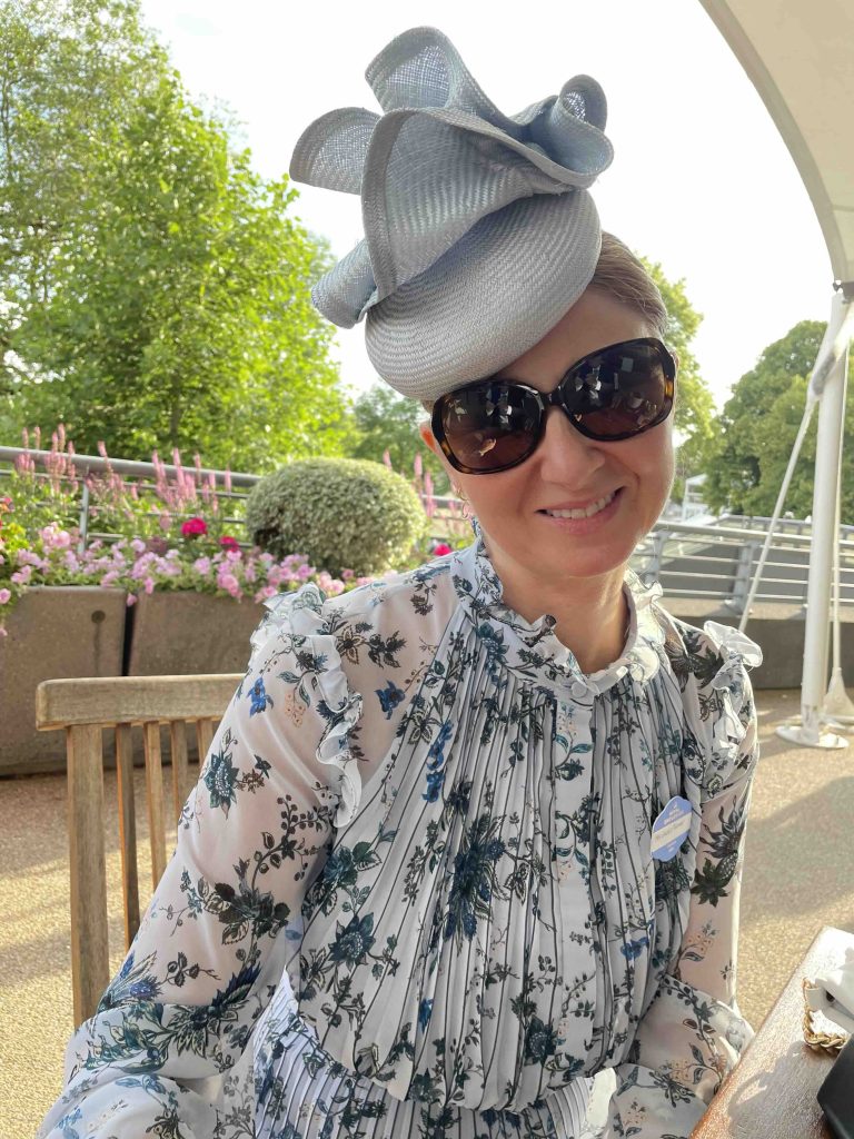 A woman in sunglasses and a floral dress smiles while wearing a large, decorative hat. She is seated outdoors with flowers and greenery in the background. | Sarah Ross Millinery