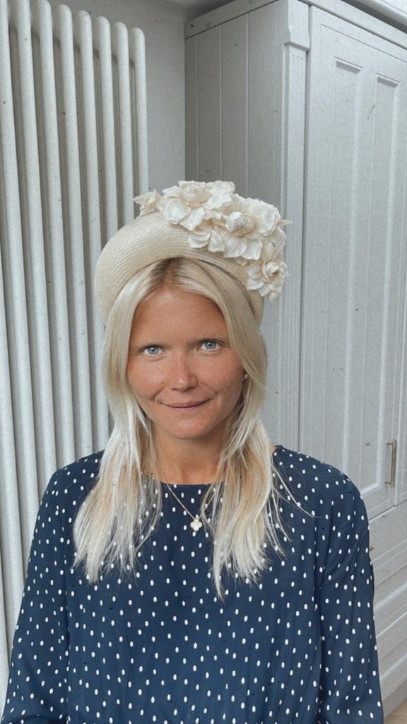 A woman with long blonde hair wears a polka dot dress and a hat adorned with white flowers, standing indoors. | Sarah Ross Millinery