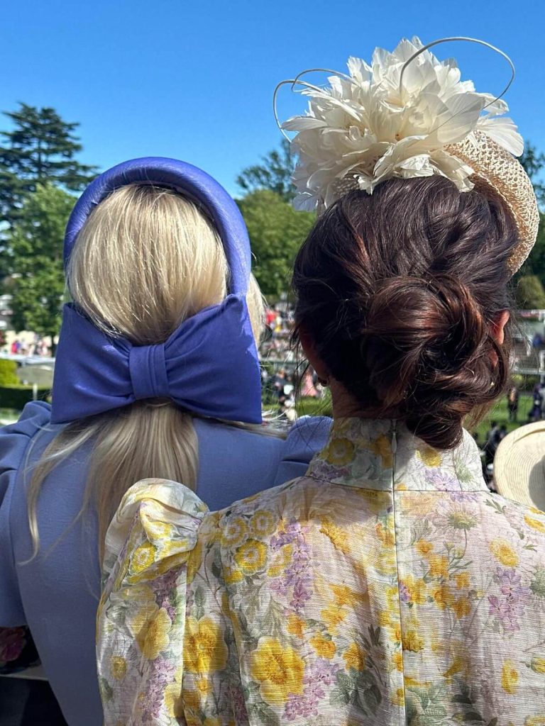 Two women standing with elaborate hairstyles and hats, dressed in floral and blue outfits, at an outdoor event on a sunny day. | Sarah Ross Millinery