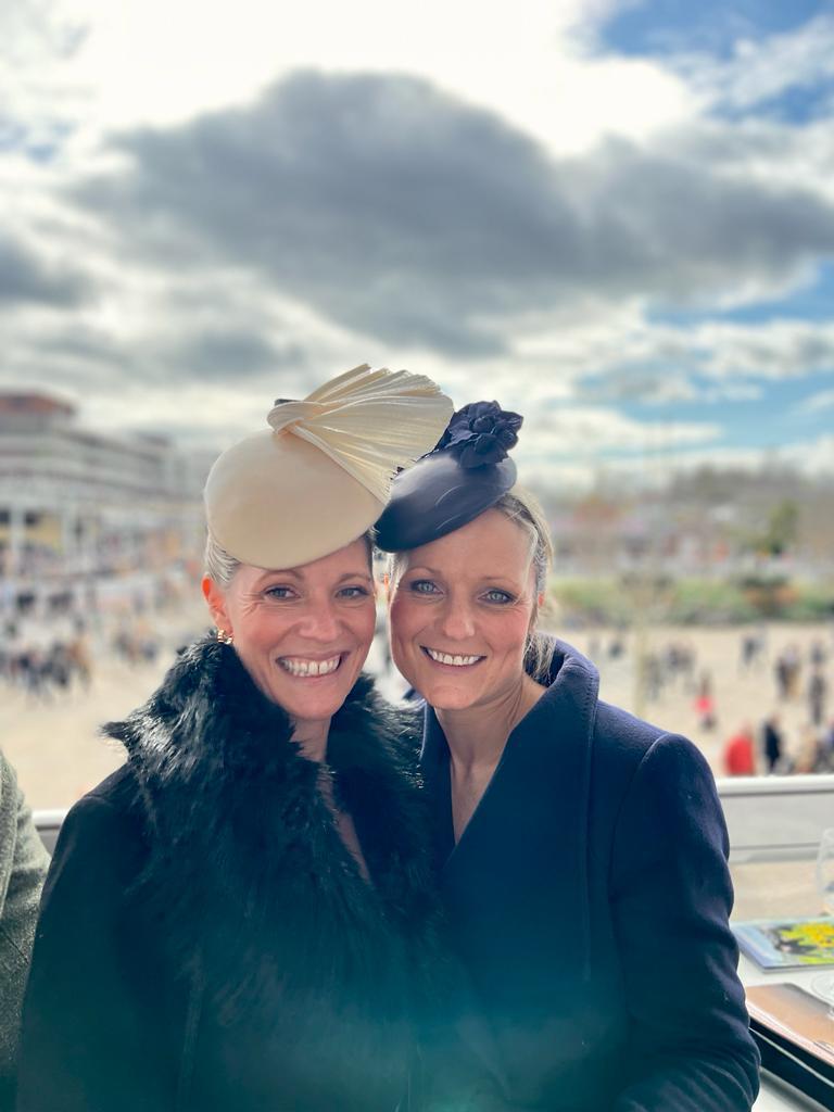 Two women wearing fashionable hats and dark coats pose together outdoors, smiling with a cloudy sky in the background. | Sarah Ross Millinery