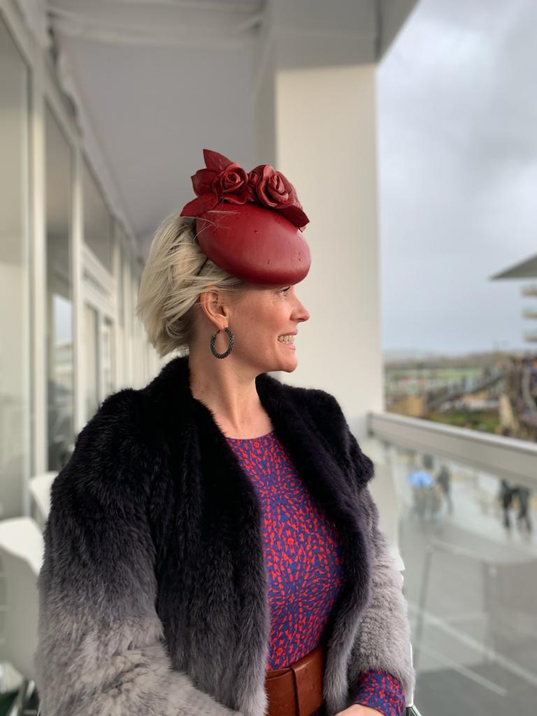 A woman with short blonde hair stands on a balcony, wearing a red hat with flowers, black fur coat, and a blue and red patterned dress. | Sarah Ross Millinery
